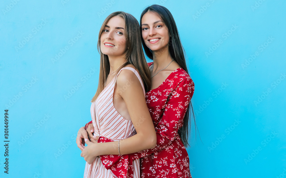 Friendly Tenderness. Close up photo of two sisters in vivid summer dresses posing in profile with their heads turned towards the camera, smiling and hugging. Friendly Tenderness. Close up photo of two sisters in vivid summer dresses posing in profile with their heads turned towards the camera, smiling and hugging.