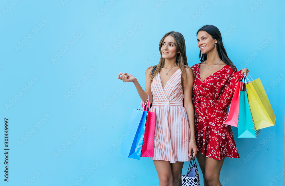Excellence shopping. A brunette and a blonde are posing together in beautiful dresses, holding a bunch of shopping bags, smiling and looking aside.