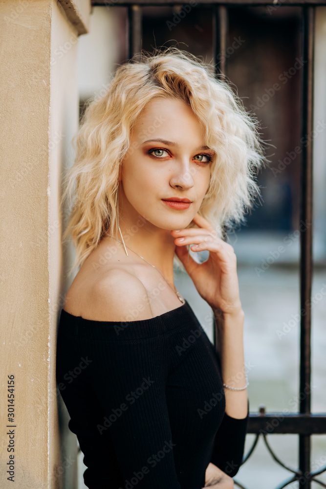 Fototapeta premium Incredible blonde woman with short haircut in black dress is leaning against wall