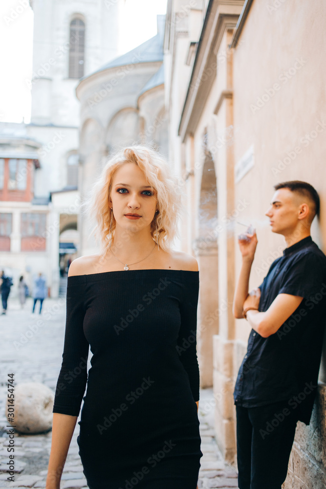 A attractive blonde in a black dress walks past a guy who smokes near a wall