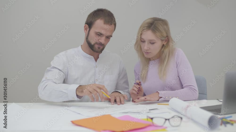 Two professional Caucasian designers looking through blueprints in the office. Young pretty woman and handsome man implementing ideas on paper. Two professional Caucasian designers looking through blueprints in the office. Young pretty woman and handsome man implementing ideas on paper.