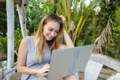 Happy blonde using laptop and sitting in exotic garedn with palms in background. photo