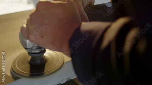 Man's hand uses electric craft polisher to professionally polish surface of grey granite block which is placed on wooden table in workshop.