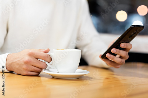 Handsome young caucasian man sitting at cafe using mobile phone and having coffee. Caucasian male with cup of coffee and reading text message on his smart phone .