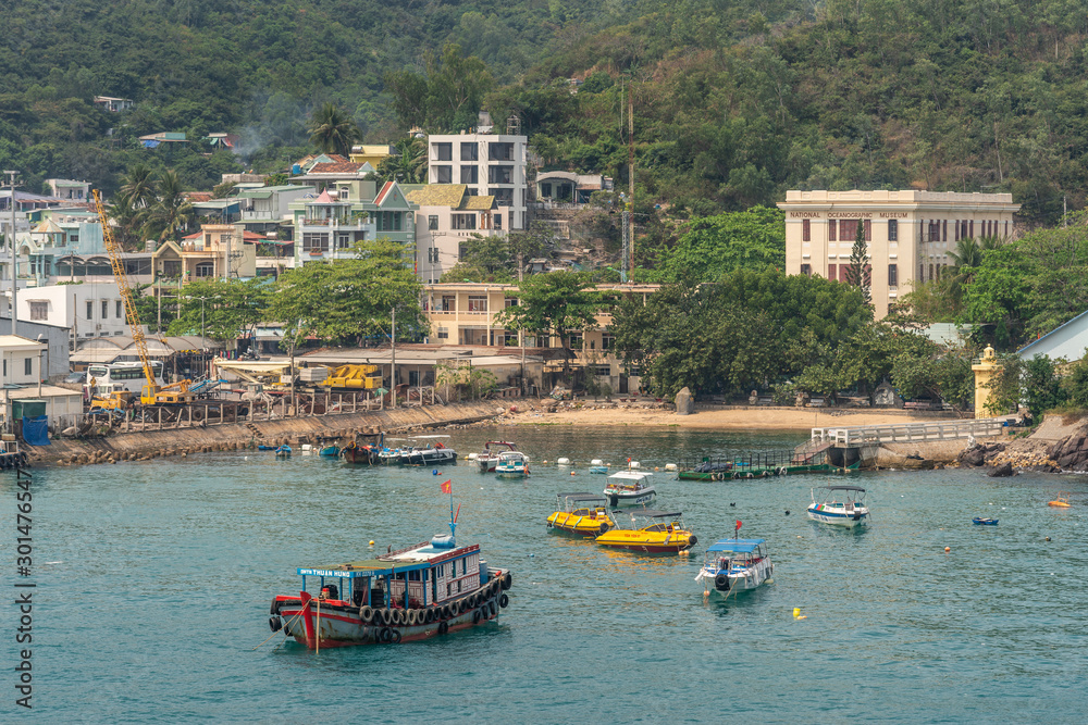 Nha Trang, Vietnam - March 11, 2019: Ferry landing pier with colorful small boats in marina adjacent to commercial port. Hotels and housing against green hill flank. 