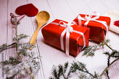 Two red Christmas boxes with white ribbon, a wooden spoon, a red heart decoration and green sprouce on a white wooden table.  photo