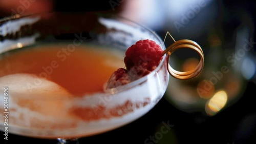 close hand held shot with slow focus shift of an orange cocktail with raspberry on a golden stirer in a coupe glass in a dark moody night surrounding bar atmosphere. Packshot with copy space. photo