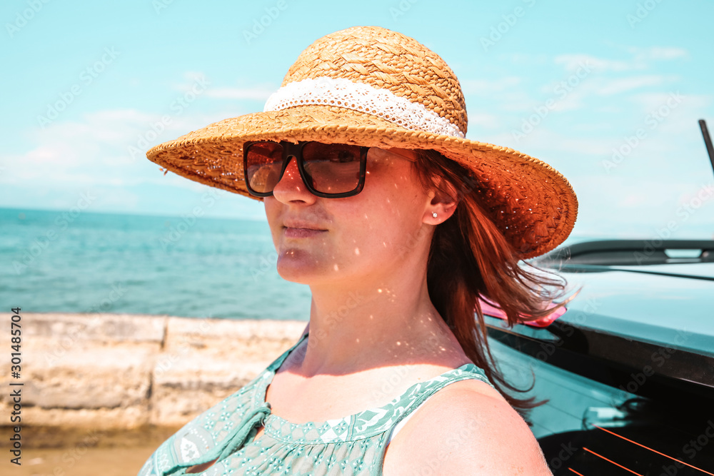 Young woman in a straw hat and sunglasses. Dark soul car on the beach. View of beautiful sea and blue sky. Place for your text or product. A beautiful summer day. Car trip and sightseeing of the coast