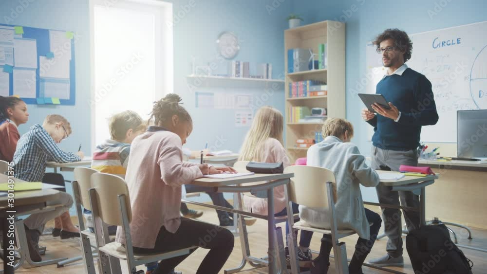 Enthusiastic Teacher Uses Digital Tablet Computer While Explaining a Lesson to a Classroom Full of Diverse Bright Children. In Elementary School Group of Smart Multiethnic Kids Learning