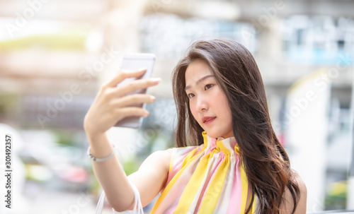 Young beautiful Asian woman holding her smartphone to taking a selfie with her shopping bags