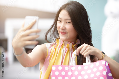 Young beautiful Asian woman holding her smartphone to taking a selfie with her shopping bags 