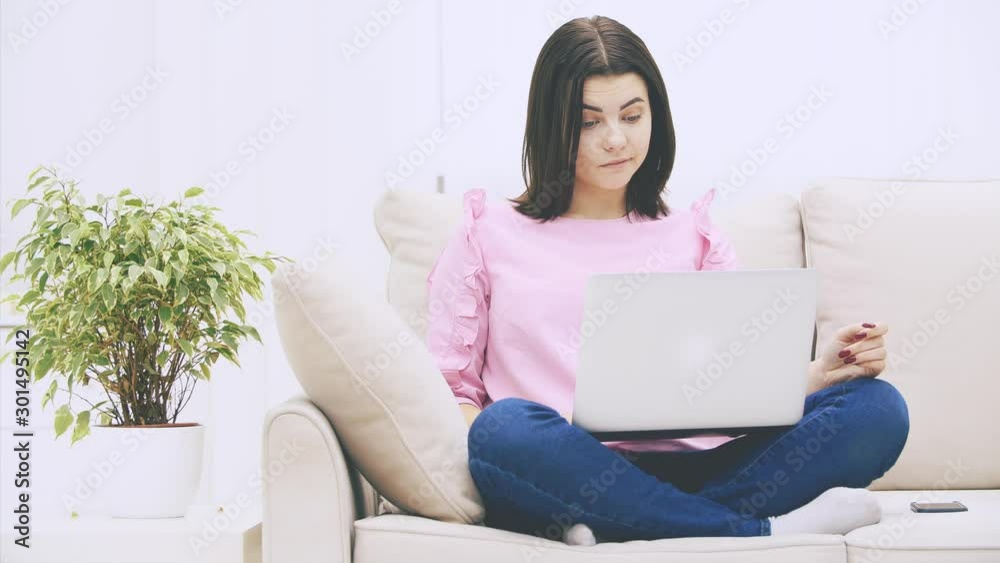 Cute caucasian brunette girl sitting on the sofa in lotos position, with laptop, surfing in the internet, throwing hands, raising eyebrows, looking confused. Cute caucasian brunette girl sitting on the sofa in lotos position, with laptop, surfing in the internet, throwing hands, raising eyebrows, looking confused.