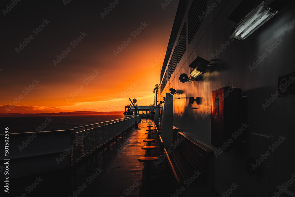 Deck of ferry sailing across the sea during last moments of a beautiful sunset with arriving land in the background. Concept of relaxation, adventure, freedom, luxury and leisure.