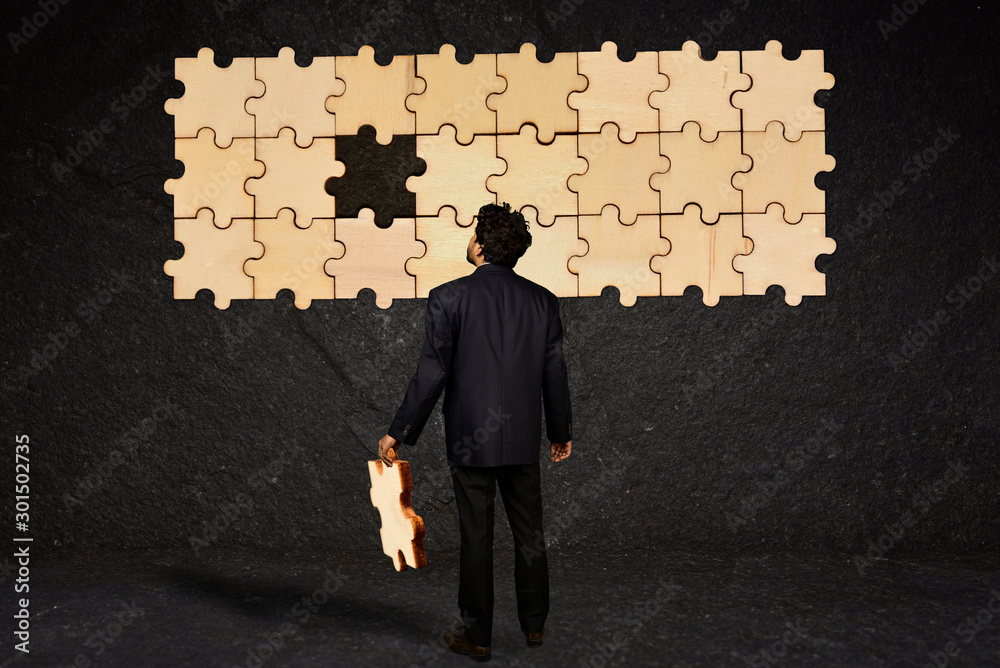 A businessman holding the missing piece of puzzle to an unfinished puzzle hanging on a black textured wall A businessman holding the missing piece of puzzle to an unfinished puzzle hanging on a black textured wall