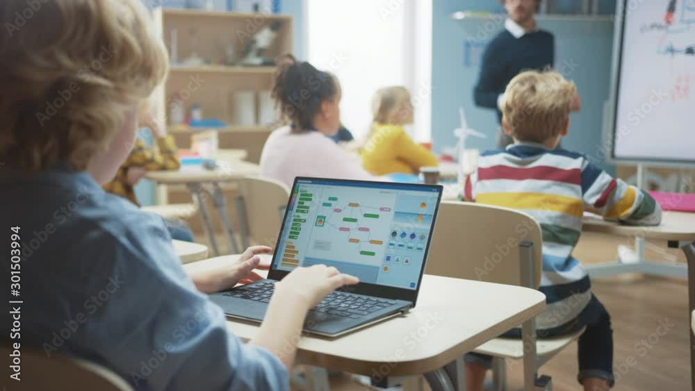 Elementary School Science Class: Over the Shoulder Little Boy Uses Laptop with Screen Showing Programming Software. Physics Teacher Explains Lesson to a Diverse Class full of Smart Kids Elementary School Science Class: Over the Shoulder Little Boy Uses Laptop with Screen Showing Programming Software. Physics Teacher Explains Lesson to a Diverse Class full of Smart Kids