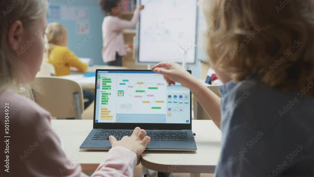 Elementary School Science Class: Over the Shoulder Little Boy and Girl Use Laptop with Screen Showing Programming Software. Physics Teacher Explains Lesson to a Diverse Class full of Smart Kids Elementary School Science Class: Over the Shoulder Little Boy and Girl Use Laptop with Screen Showing Programming Software. Physics Teacher Explains Lesson to a Diverse Class full of Smart Kids