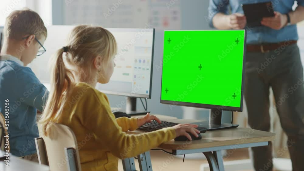 Elementary School Computer Science Classroom: Cute Little Girl Uses Green Mock-up Screen Computer while Learning Coding and Programming. Schoolchildren Getting Modern Education. Over the Shoulder