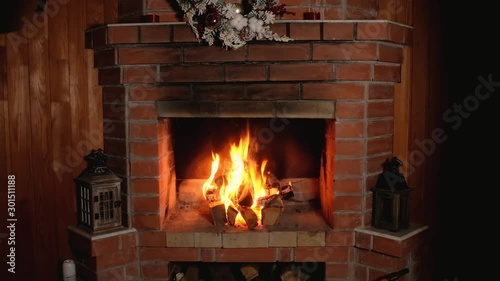 flame of fire burns in brick home Christmas fireplace. festive wreath hangs over fireplace. Wooden candlesticks is decorated with Christmas decor. Rest and relaxation in front of fireplace in evening.