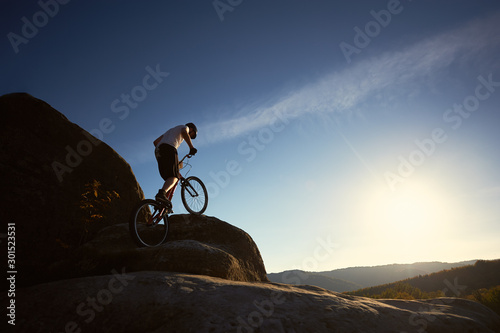 Bilde på lerret Professional cyclist balancing on trial bicycle, male rider making acrobatic stunt on top of big boulder on summer sunny day, blue sky and sunset on background