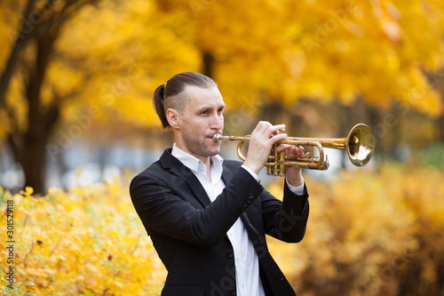 young European handsome trumpeter in formal clothes playing his musical instrument golden trumpet among trees with yellow leaves in autumn park photo