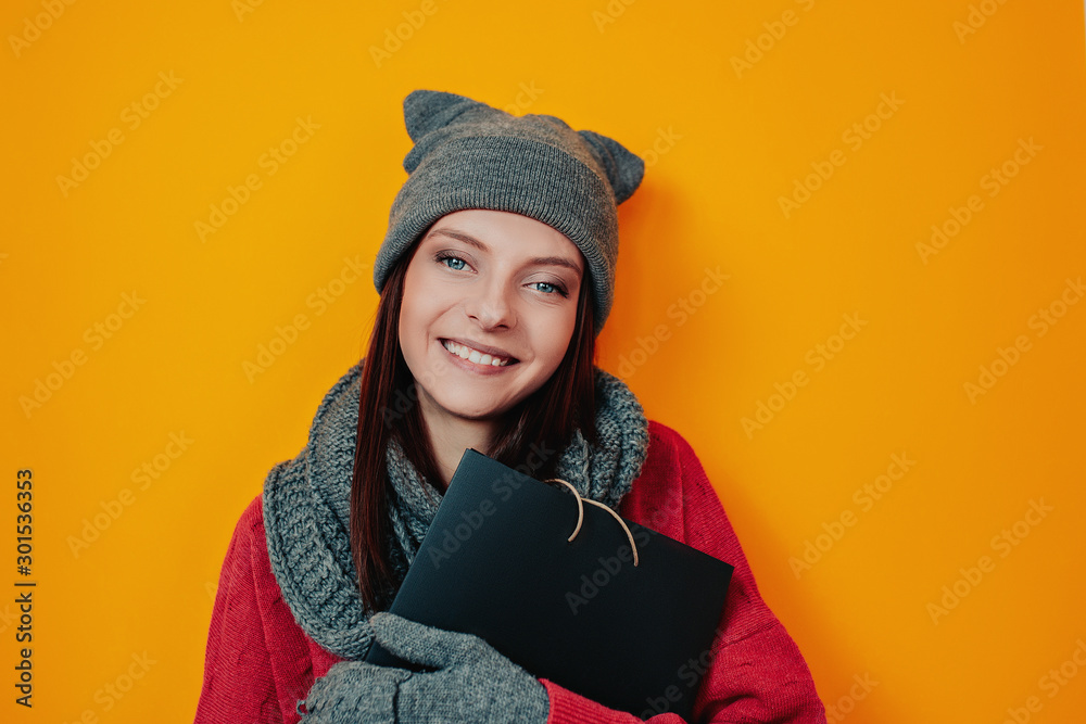 Woman in gray hat on the orange background. Young woman in sweater and hat holds black bag. Black friday image. Woman shopping. Woman in gray hat on the orange background. Young woman in sweater and hat holds black bag. Black friday image. Woman shopping.