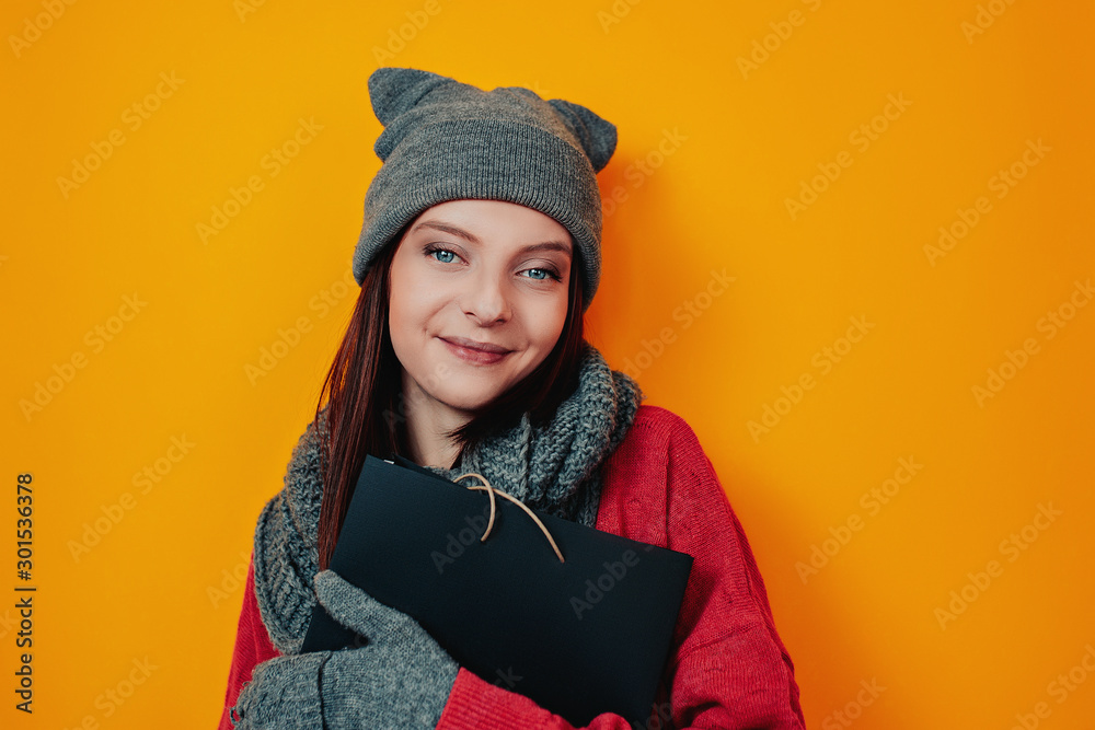 Woman in gray hat on the orange background. Young woman in sweater and hat holds black bag. Black friday image. Woman shopping. Woman in gray hat on the orange background. Young woman in sweater and hat holds black bag. Black friday image. Woman shopping.