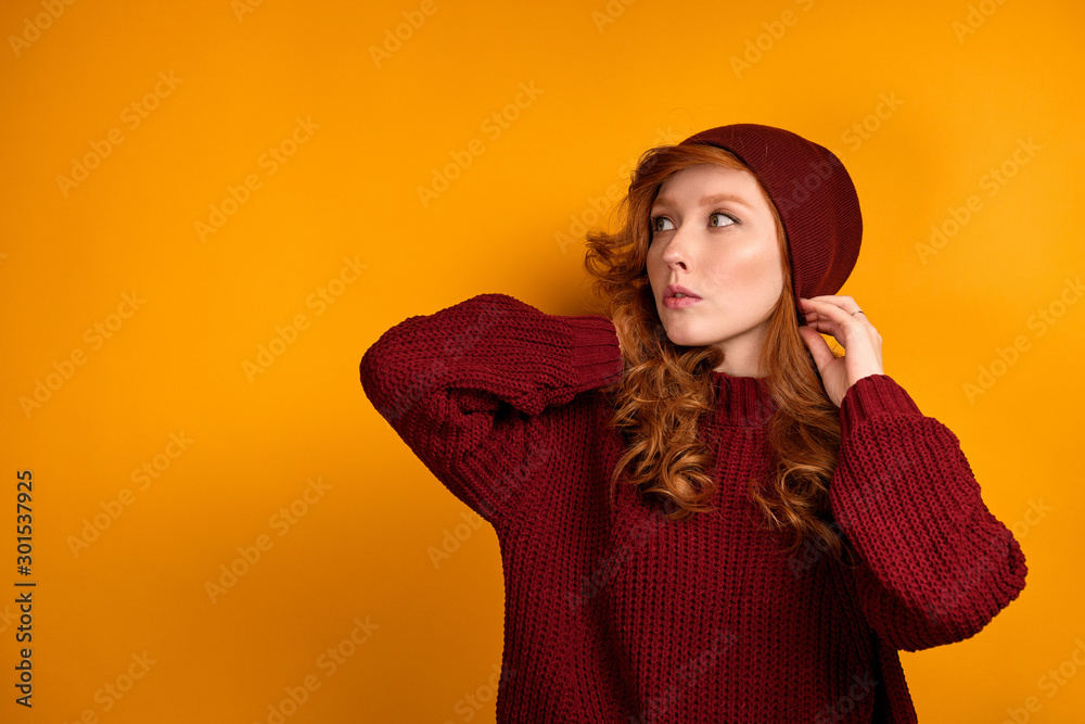 A curly red-haired girl in a burgundy sweater and hat stands on an orange background, straightening her hat, looks to the side. A curly red-haired girl in a burgundy sweater and hat stands on an orange background, straightening her hat, looks to the side.