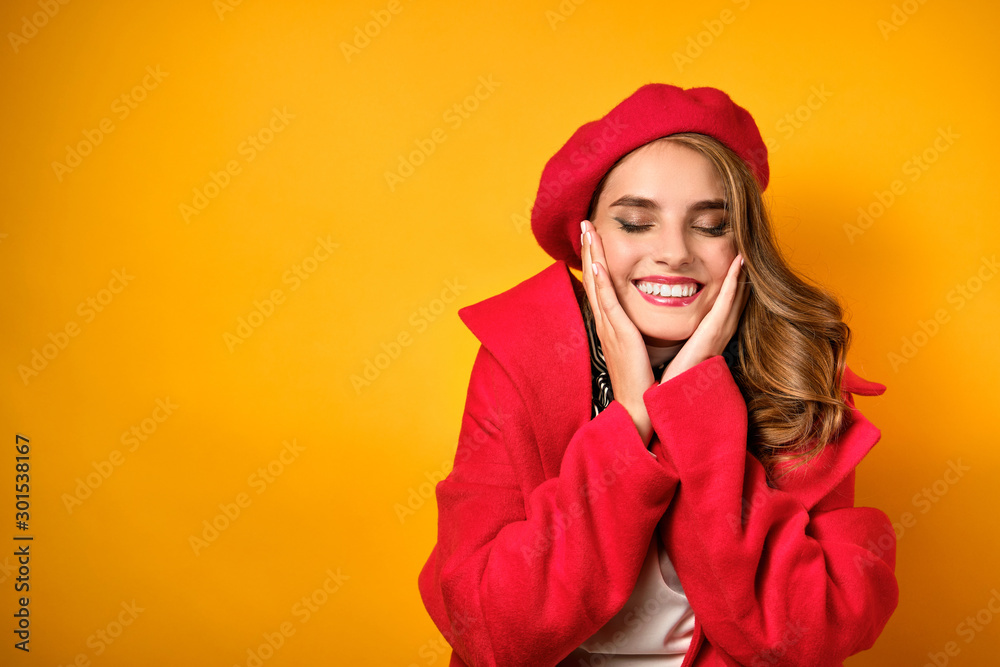 A girl with red lipstick in a red coat and beret is standing on a yellow background, squinting pretty, presses palms to her face A girl with red lipstick in a red coat and beret is standing on a yellow background, squinting pretty, presses palms to her face