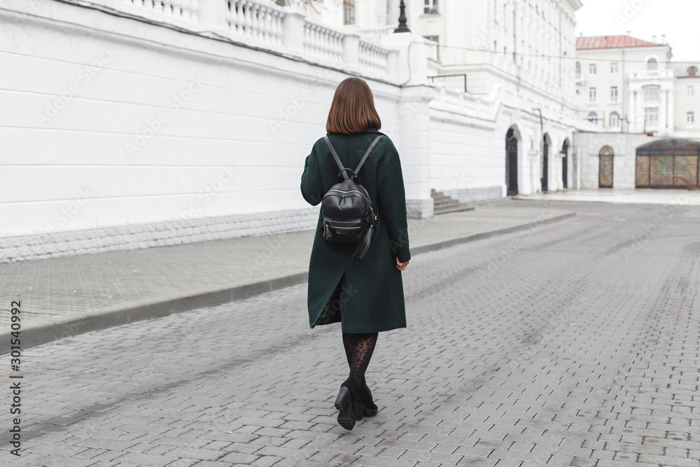 Beautiful young stylish brunette woman wearing midi green coat, black heeled boots, backpack, leopard print tights walking through the city streets. Trendy casual outfit. Street fashion. Back view.