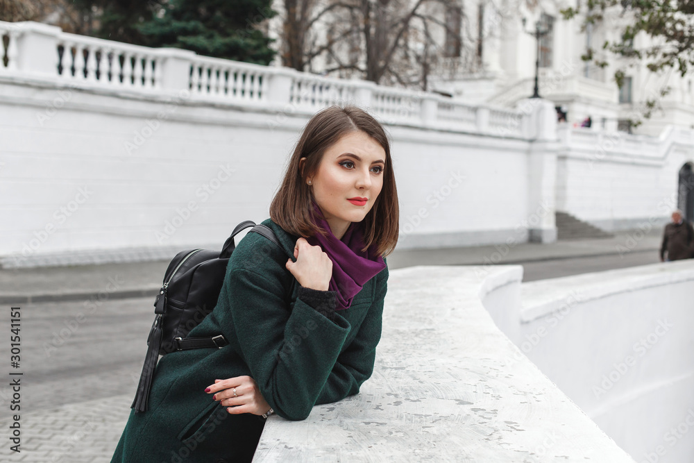 Beautiful young stylish brunette woman with short haircut, wearing green coat, black backpack, purple scarf walking through the city streets. Trendy casual outfit. Everyday look. Street fashion.