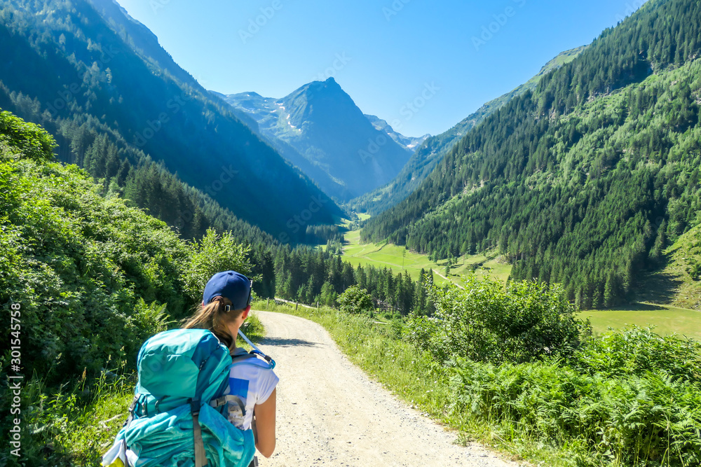 A young woman with a big hiking backpack walking on a wide pathway in a valley. Beauty of the nature. Lush green grass and trees overgrowing the meadow. Spring in the Alps.
