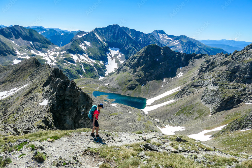 A young woman with a big backpack hikes down towards a clear, navy blue lake hiding between tall mountain peaks. Some of the slopes are covered with snow. In the back is another mountain range