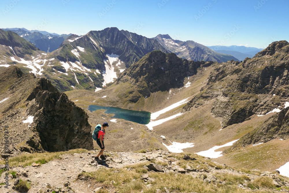 A young woman with a big backpack hikes down towards a clear, navy blue lake hiding between tall mountain peaks. Some of the slopes are covered with snow. In the back is another mountain range