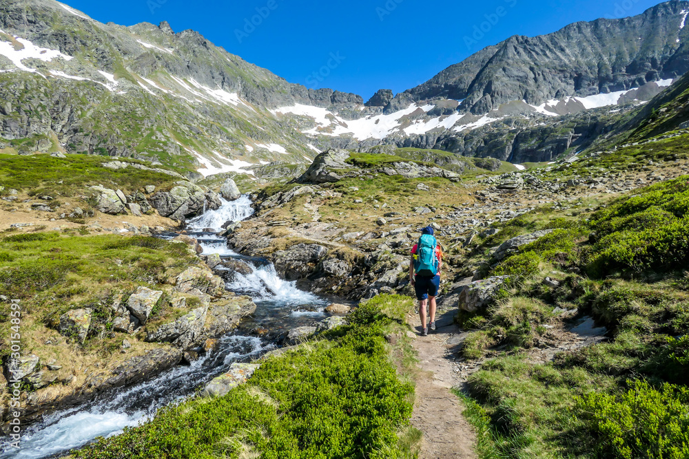 A young woman with a big backpack hikes up on a steep pathway, along a mountain stream. the water cascades on the rocks, rushing into the valley. Girl is enjoying the spring in the mountains