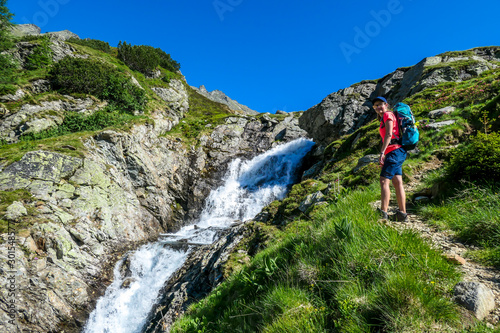 A young woman with a big backpack hikes up on a steep pathway, along a mountain stream. the water cascades on the rocks, rushing into the valley. Girl is enjoying the spring in the mountains