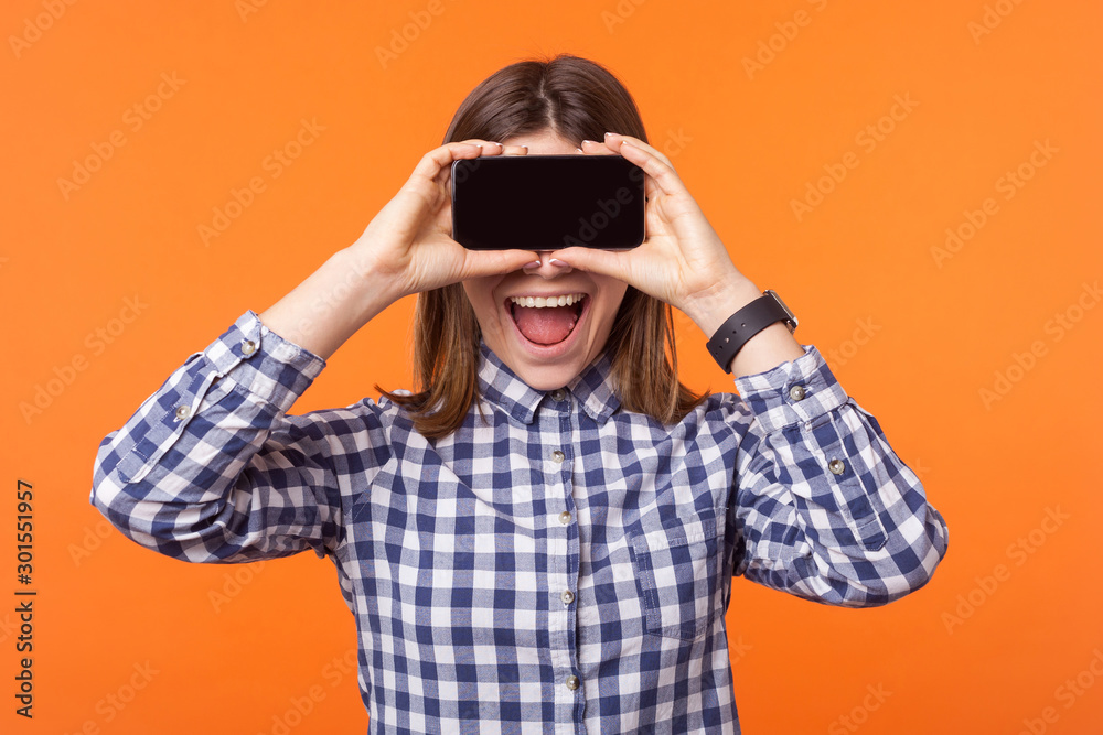 Portrait of happy amazed woman with toothy smile and brown hair wearing checkered shirt standing, hiding her eyes with cellphone, taking pictures. indoor studio shot isolated on orange background Portrait of happy amazed woman with toothy smile and brown hair wearing checkered shirt standing, hiding her eyes with cellphone, taking pictures. indoor studio shot isolated on orange background