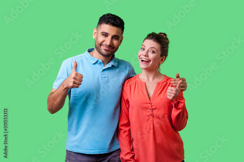 Portrait of positive attractive young couple in casual wear standing together, hugging as friends, showing thumbs up gesture and smiling at camera. isolated on green background, indoor studio shot