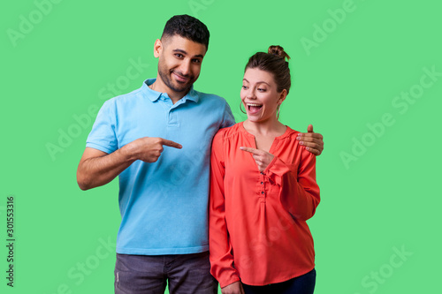 Portrait of cheerful attractive young couple in casual wear standing together, hugging as friends, pointing fingers and choosing each other, smiling. isolated on green background, indoor studio shot