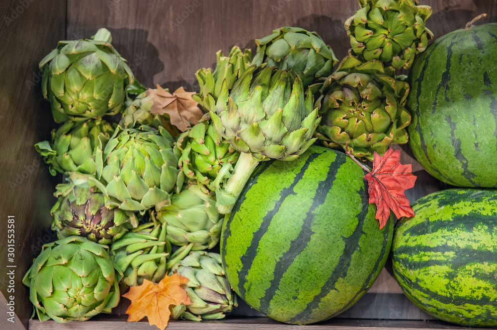 Obraz premium Farm vegetables ripe watermelons and artichokes lie in a box in the window of the farmers market. The latest varieties and hybrids of fruits and vegetables