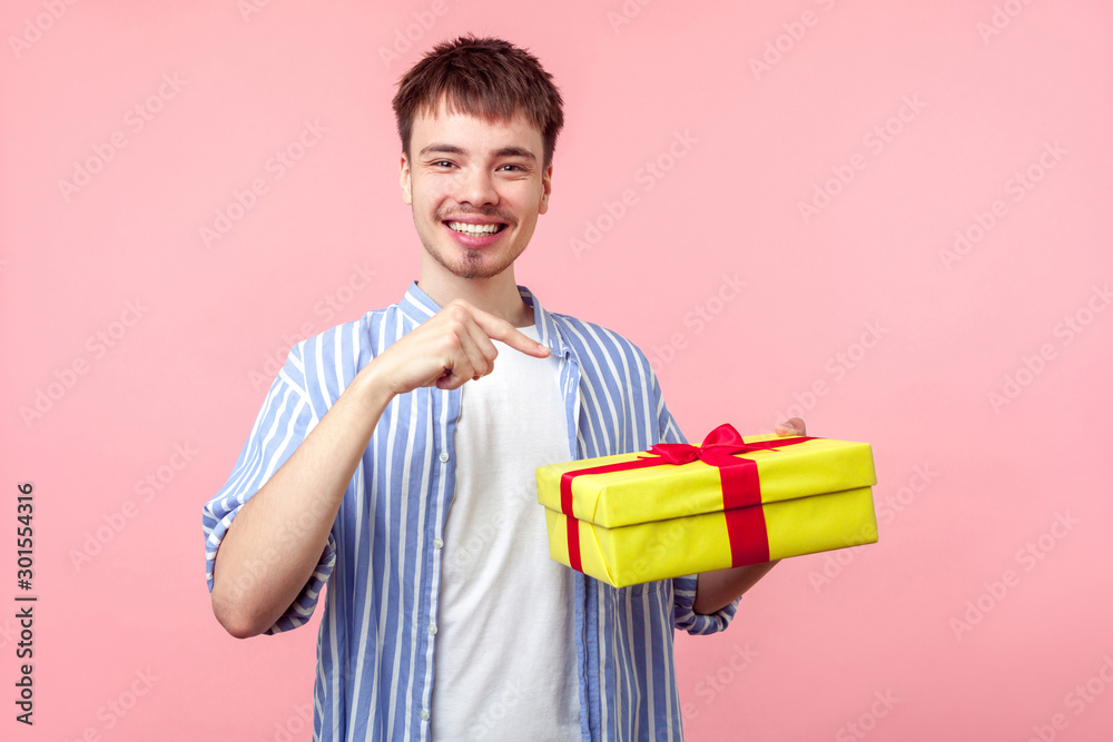 Portrait of happy young brown-haired man with small beard and mustache in casual shirt pointing at gift box and smiling at camera, enjoying present. indoor studio shot isolated on pink background Portrait of happy young brown-haired man with small beard and mustache in casual shirt pointing at gift box and smiling at camera, enjoying present. indoor studio shot isolated on pink background