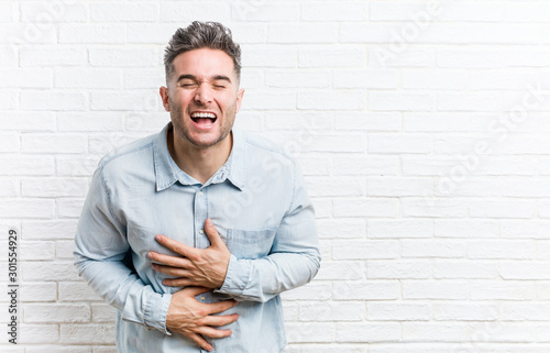 Young handsome man against a bricks wall laughs happily and has fun keeping hands on stomach.