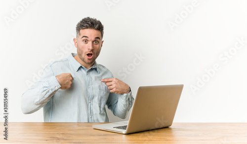 Young handsome man working with his laptop surprised pointing with finger, smiling broadly.