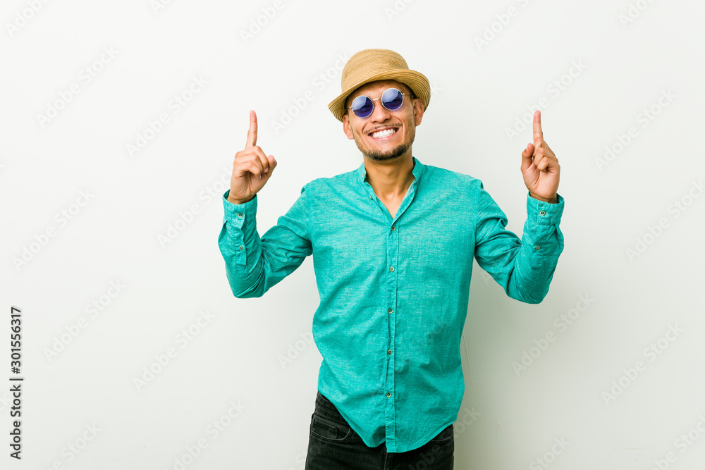 Young hispanic man wearing a summer clothes indicates with both fore fingers up showing a blank space.
