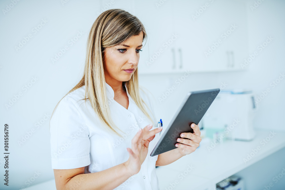 Portrait of beautiful blond female lab assistant in white uniform using tablet while standing in laboratory. Portrait of beautiful blond female lab assistant in white uniform using tablet while standing in laboratory.