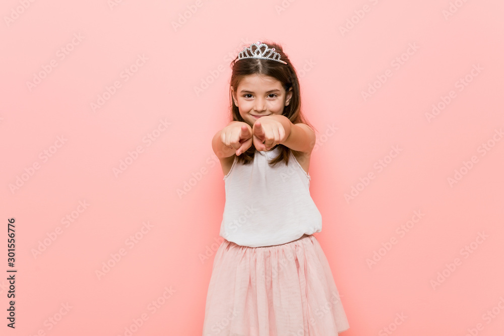 Little girl wearing a princess look cheerful smiles pointing to front. - obrazy, fototapety, plakaty 