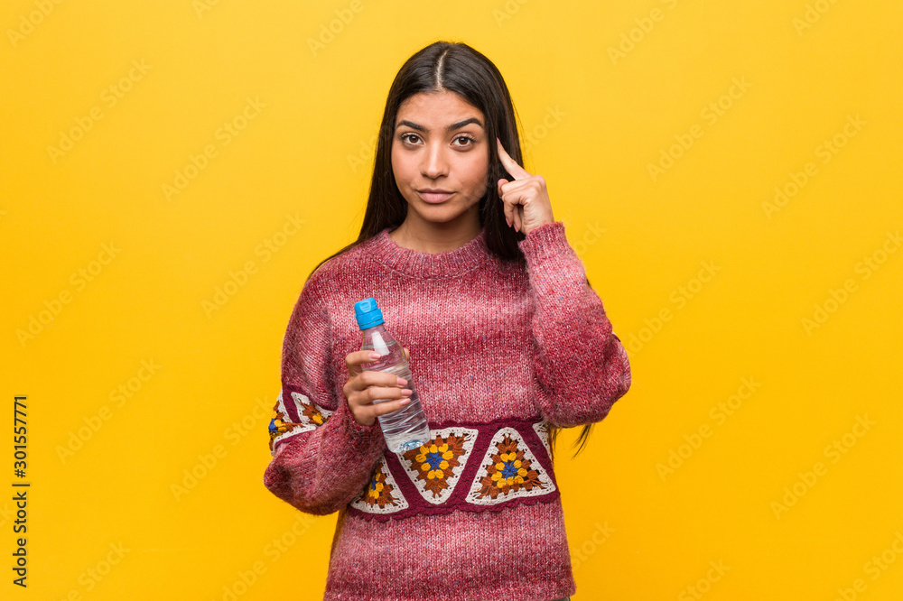 Young arab woman holding a water bottle pointing his temple with finger, thinking, focused on a task. Young arab woman holding a water bottle pointing his temple with finger, thinking, focused on a task.