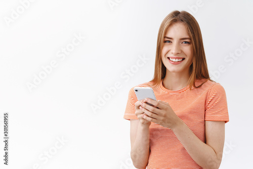 Friendly, pretty young carefree woman in striped t-shirt hold smartphone, smiling and gazing camera delighted, browsing internet, making phone call, texting funny story, stand white background