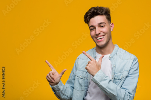 Young handsome student wearing a denim shirt pointing with forefingers to a copy space, expressing excitement and desire.