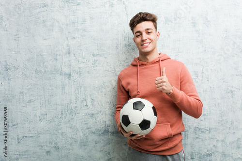 Young sporty man holding a soccer ball smiling and raising thumb up