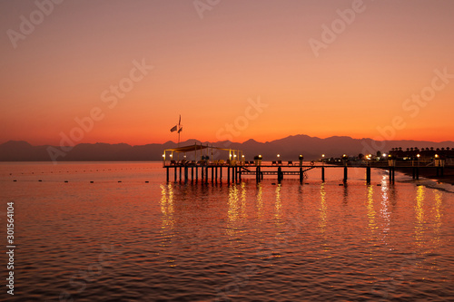 Beautiful golden sunset sea landscape with wooden pier of hotel resort  shiny sea water with reflected lights on surface and silhouettes of dark mountains at horizon. Horizontal color photography.